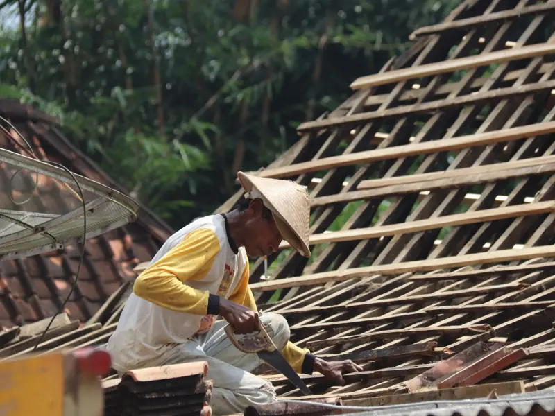 Carpenter working on a wooden roof structure outdoors in a rural setting with traditional tools.