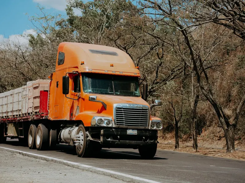 A vibrant orange Freightliner truck drives on a highway through a dry, scenic landscape in El Salvador.