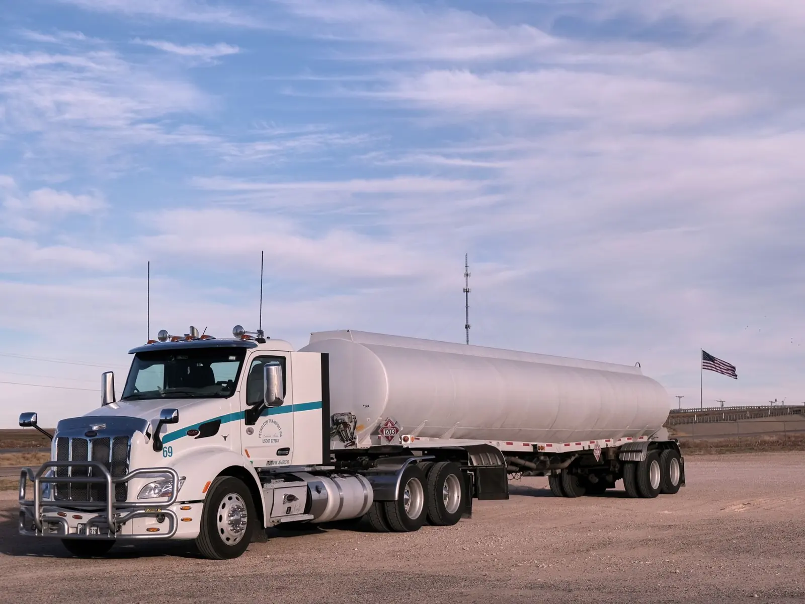 White fuel transport truck parked in a Texas landscape under clear skies, showcasing American flag in the distance.