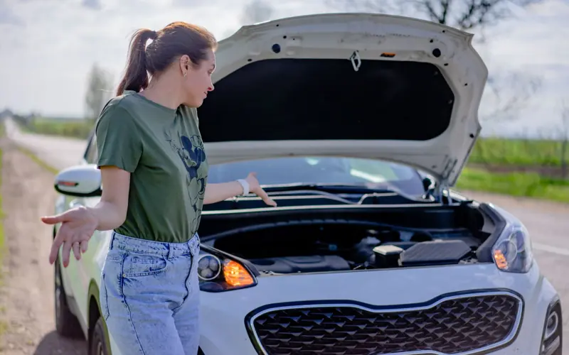 A woman looks puzzled by her broken car with an open hood on a rural road.