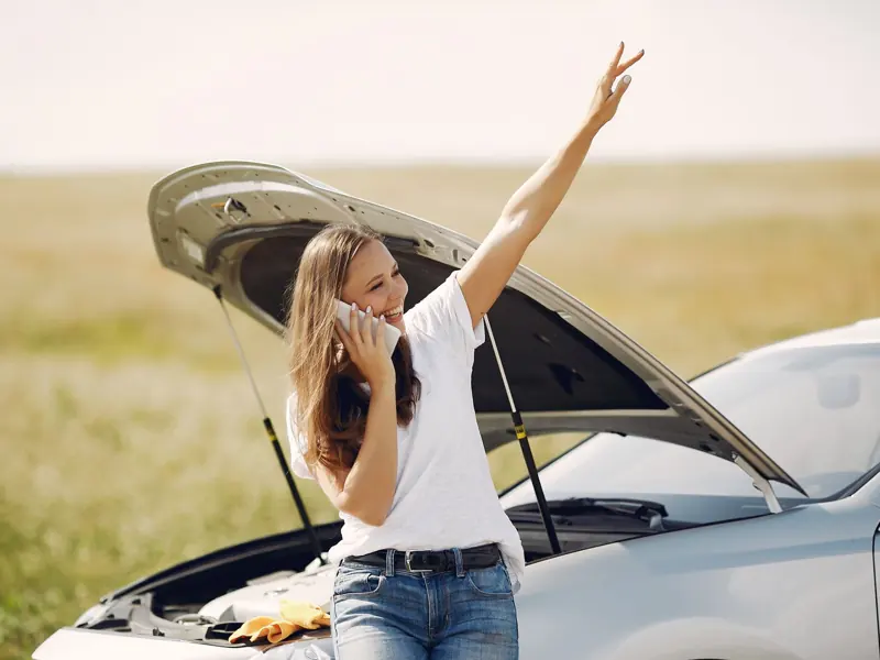 Cheerful young female driver in white t shirt and jeans speaking on phone and looking away with smile while leaning on broken car with open hood and showing greeting gesture with raised hand