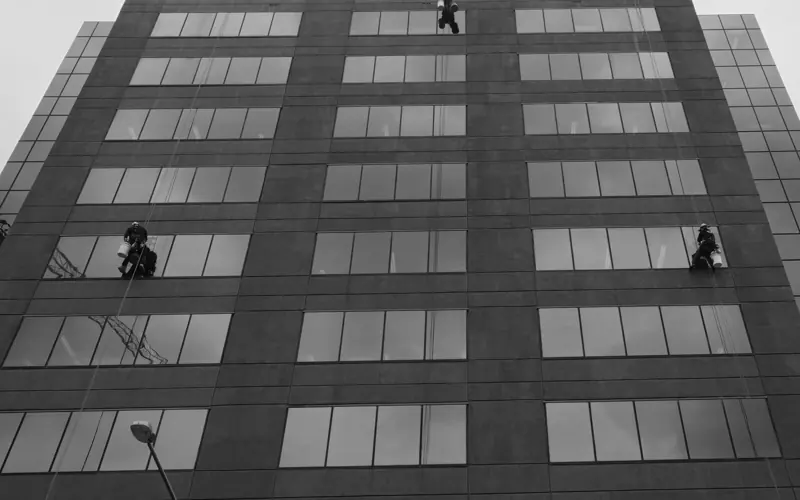 Window cleaners on skyscraper facade performing maintenance duties in monochrome setting.