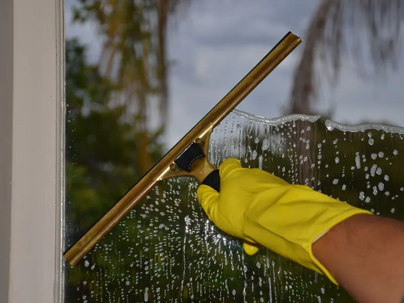 Close-up of hand cleaning window with squeegee outdoors using yellow gloves.